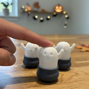 Puede incluir: Tres figuras blancas con forma de fantasma con los brazos extendidos y expresiones de sorpresa. Cada fantasma está sentado en una base gris oscuro. Las figuras están sobre una superficie de madera, con decoraciones de Halloween en el fondo.