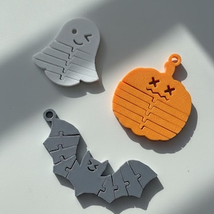 Puede incluir: Tres piezas de rompecabezas con temática de Halloween: un fantasma blanco sonriente, una calabaza naranja con ojos de cruz y un murciélago gris. Cada pieza tiene un lazo para colgar y está hecha de un material suave.