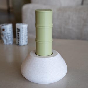 Peut inclure: Un objet cylindrique vert clair avec trois crêtes horizontales, ressemblant à une tige de bambou, est inséré dans une base blanche en forme de dôme avec une texture granuleuse rappelant la pierre. La base présente une ouverture circulaire.