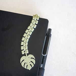 May include: A light green, articulated, segmented bookmark featuring a monstera leaf design at the base, resting on a black notebook next to a black pen with the text 3D printed on the side of the pen barrel near the grip area.
