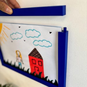 May include: A blue picture frame holding a child's drawing of a house, clouds, and a person. The frame is being held up against a white wall. The drawing is in bright colors.