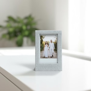 May include: A light gray, textured picture frame with a photograph of a couple holding hands. The frame is rectangular with a slightly raised border. The photo shows a bride and groom walking on a path.