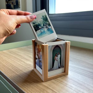 Puede incluir: Un cubo de fotos de madera marrón claro que muestra varias fotos instantáneas. El cubo contiene una pila de fotos instantáneas, una de las cuales se levanta para revelar las demás. Las fotos muestran varias escenas, incluyendo una boda y una fiesta en la piscina.