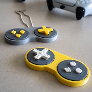 May include: Close-up of two decorative video game controller keychains. One is gray and yellow, the other is gray with yellow accents. Two full-size controllers are in the background. The keychains have a retro design.