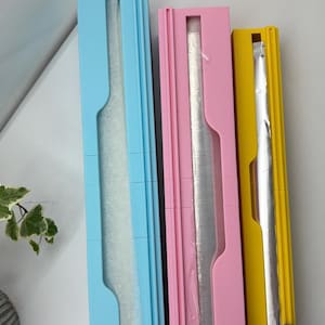 May include: Three plastic wrap dispensers in different colours: light blue, pink, and yellow. Each dispenser has a clear plastic window to see the wrap inside.