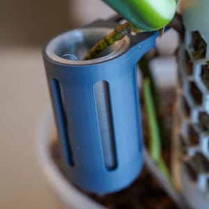 May include: A matte blue cylindrical plant propagation vessel with vertical cutouts, featuring a translucent inner sleeve. The vessel is attached to a white hexagonal grid structure, supporting a green plant stem extending into the open top.