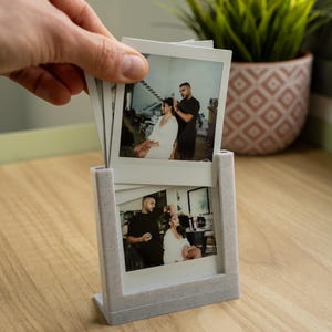 May include: A light gray photo frame holding several instant photos. The photos depict a person getting their hair styled. The frame is on a wooden surface. A hand is holding more photos above the frame. A potted plant is in the background.