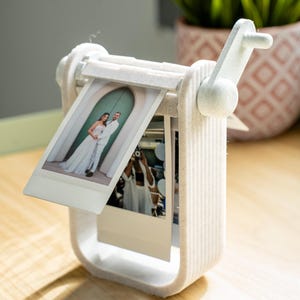 Könnte beinhalten: Ein weißer, 3D-gedruckter Foto-Display mit Kurbel, der zwei Sofortbilder hält. Die Fotos zeigen ein Hochzeitspaar und eine andere Person, die ein Foto macht. Das Display steht auf einer Holzoberfläche, mit einer Pflanze im Hintergrund.