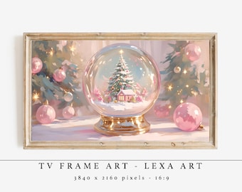 Op de afbeelding: Een ingelijste digitale kunstprint met een sneeuwbol met een kerstboom en een klein huisje erin. Het kunstwerk heeft een zacht, pastelkleurenpalet met roze en gouden accenten. De lijst is licht van kleur. De tekst onderaan luidt "TV FRAME ART - LEXA ART".