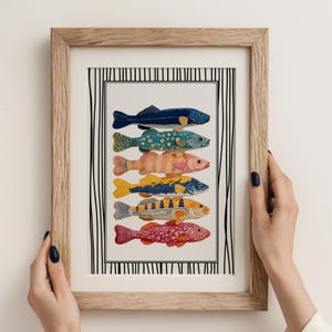May include: A framed art print displaying six vibrantly coloured fish illustrations. The fish are in various colours and patterns, including blue, green, yellow, and red. The artwork is set against a white background with a black and white striped border, framed in light wood.