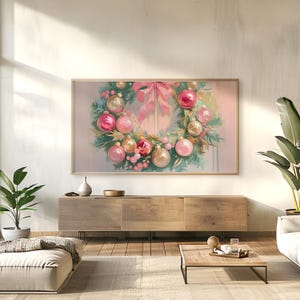 Op de afbeelding: Een ingelijst schilderij van een kerstkrans. De krans heeft roze en gouden ballen, roze bessen en een grote roze strik. De krans staat tegen een zachte, lichte achtergrond.