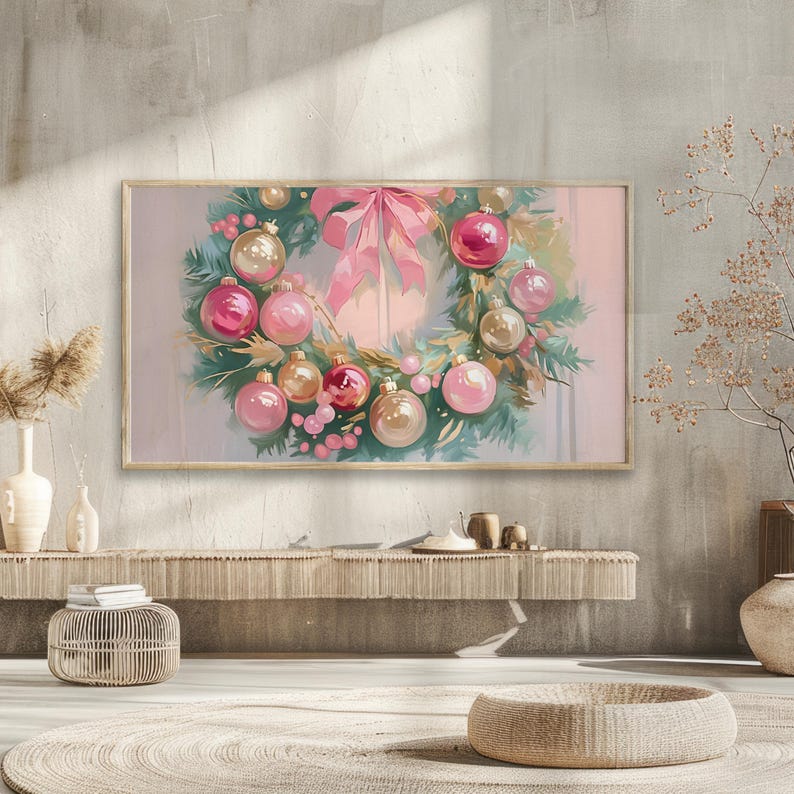 Op de afbeelding: Een ingelijste aquarel van een kerstkrans. De krans heeft roze en gouden ballen, roze bessen en een grote roze strik. De krans staat tegen een zachte, pastelkleurige achtergrond.