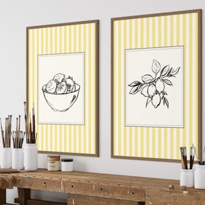 Peut inclure: Deux impressions encadrées avec des fonds à rayures jaunes et blanches. L'une représente un bol de citrons, l'autre une branche de citron. Les cadres sont bruns. Les impressions sont affichées au-dessus d'un établi en bois avec des fournitures d'art.