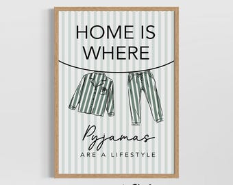 May include: A framed print with the text "HOME IS WHERE Pyjamas ARE A LIFESTYLE". The artwork features a sketch of a green and white striped pajama set hanging on a clothesline against a striped background. The frame is a light brown color.