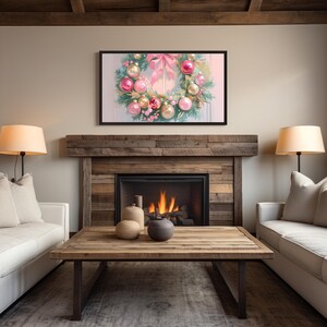 Op de afbeelding: Een ingelijst kunstwerk met een kerstkrans met roze en gouden ornamenten en een roze strik. Het kunstwerk hangt boven een open haard met een brandend vuur. De kamer heeft een rustieke houten salontafel, twee lampen en twee witte banken.