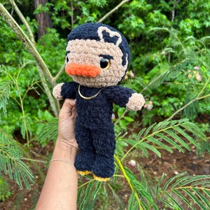 May include: A handmade crochet doll with a black outfit, tan face, and orange beak. The doll has a heart-shaped design on its head and a gold chain necklace. The doll's feet have yellow soles. The doll is held in front of a green leafy background.