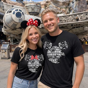 May include: Two black short-sleeved t-shirts featuring a white graphic of a Death Star shaped like Mickey Mouse ears. The left shirt includes a red and white polka dot bow graphic. The shirts are worn with blue jeans and khaki pants.