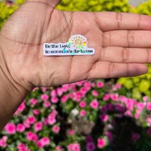 May include: A white sticker with a gold sun and the text "Be the light in someone's darkness".
