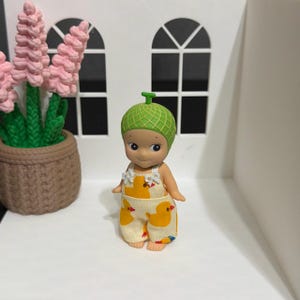 May include: A small figurine wearing a green melon-shaped hat and cream-colored overalls featuring a yellow duck pattern. The figurine has a smooth, light-toned complexion, dark eyes, and a small smile, standing upright with arms at its sides.