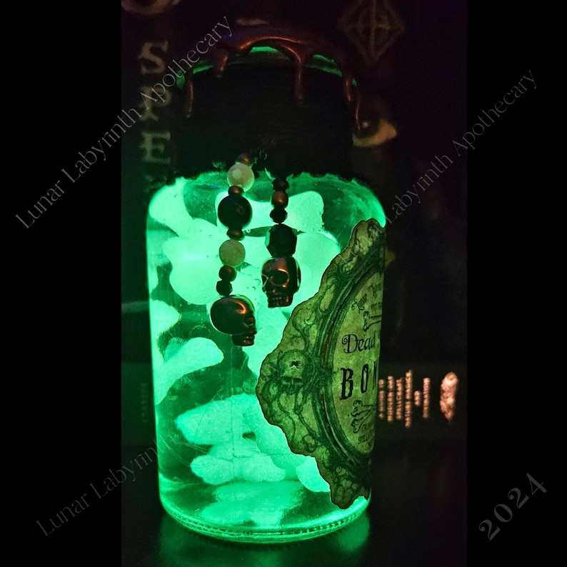 Op de afbeelding: Een glazen pot met een groene gloed van binnen. De pot heeft een zwart deksel met druppelend was en een vintage label met de tekst "Dead Bones". De pot is versierd met een kralen schedelbedel.