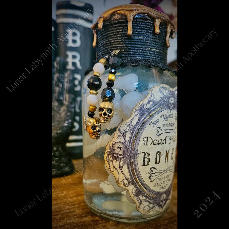 Op de afbeelding: Een glazen pot met een zwart en gouden deksel gevuld met witte stenen en een schedelbedel. De pot heeft een vintage label met de tekst "Dead Man's Bones" en "Crush & Burn Spells in My Lunar Labyrinth Apothecary".