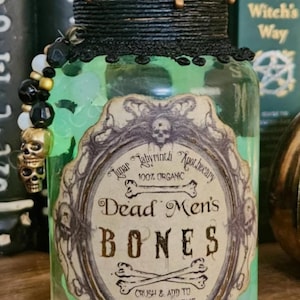 Op de afbeelding: Een groene glazen fles met een gouden dop en een zwart etiket met de tekst "Lunar Labyrinth Apothecary 100% Organic Dead Men's Bones Crush & Add To Spells To Increase Longevity".