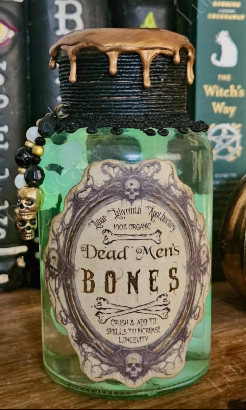 Op de afbeelding: Een groene glazen fles met een gouden dop en een zwart etiket met de tekst "Lunar Labyrinth Apothecary 100% Organic Dead Men's Bones Crush & Add To Spells To Increase Longevity".
