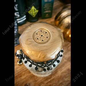 Op de afbeelding: Een gouden pot met een zwarte kanten rand en een gouden deksel met een zwart ontwerp. De pot staat op een houten oppervlak.