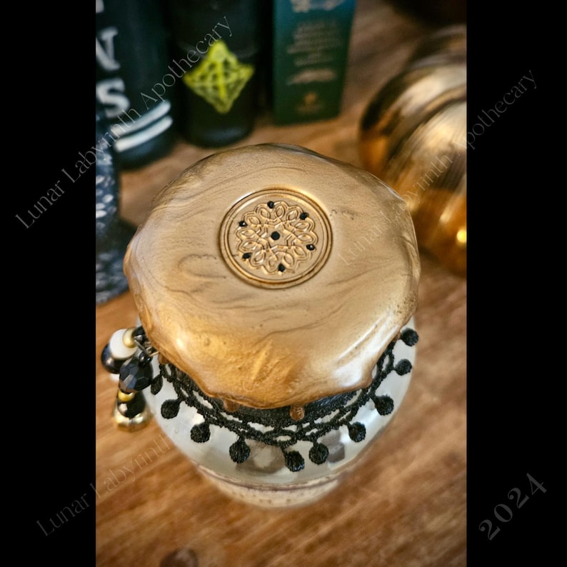 Op de afbeelding: Een gouden pot met een zwarte kanten rand en een gouden deksel met een zwart ontwerp. De pot staat op een houten oppervlak.
