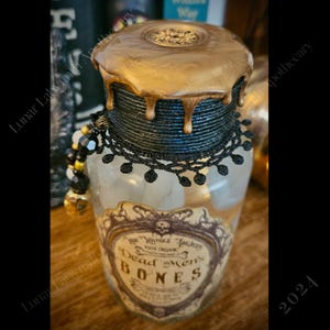 Op de afbeelding: Een glazen fles met een zwarte kanten rand en een gouden deksel. De fles is gevuld met een witte substantie en heeft een etiket met de tekst "Dead Men's Bones".