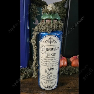 May include: A blue glass bottle labeled Gravewater Elixir with a cream-colored paper label featuring black text and a botanical illustration. The bottle neck is wrapped in a textured olive green yarn and topped with a small, sculpted skull.