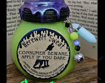 May include: A green glass jar with a black lid and a label that reads "Werewolf Sweat Consumer Beware Apply If You Dare". The jar is decorated with glitter and has a charm attached to the lid. The jar is sitting on a wooden surface.