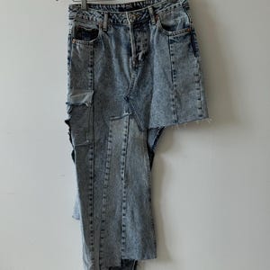 May include: Acid wash denim jeans with a unique deconstructed design. The jeans feature a skirt-like panel attached to one leg, with raw edges and visible stitching. The jeans are hanging on a wooden hanger against a white wall.