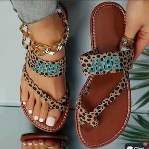 May include: A pair of brown leather sandals with a leopard print design. The sandals have a strap across the toe and a strap around the ankle. The straps are embroidered with the word "JAGS" in turquoise.