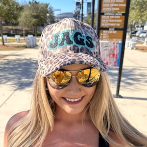 May include: A woman wearing a black tank top with a teal and gold jaguar graphic, a leopard print baseball cap with the word "JAGS" in teal and black lettering, and gold-rimmed sunglasses.
