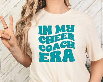 Peut inclure: T-shirt crème avec l'inscription "IN MY CHEER COACH ERA" en turquoise, dans un style rétro. Le t-shirt est noué à la taille. L'image montre une personne faisant le signe de la paix.