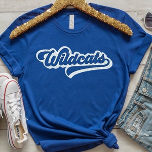 May include: Royal blue t-shirt with the word "Wildcats" in a white, retro-style font. The shirt is displayed with white sneakers, denim shorts, and heart-shaped sunglasses on a light wood surface.