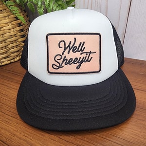 Puede incluir: Gorra de camionero blanca y negra con un parche rosa que dice "Well Sheeyit" en letras negras.