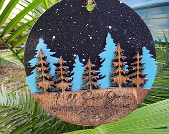 May include: Round wooden ornament with a blue and black starry night sky scene. The ornament features a cutout of a forest with pine trees and the text "I'll See You On The Other Side Of The Stars".