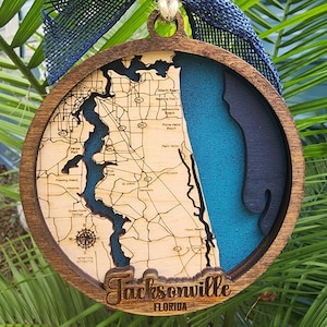 May include: A wooden ornament shaped like a map of Jacksonville, Florida. The ornament is painted in shades of blue and brown, with a blue ribbon attached to the top. The text "Jacksonville, Florida" is engraved on the bottom of the ornament.