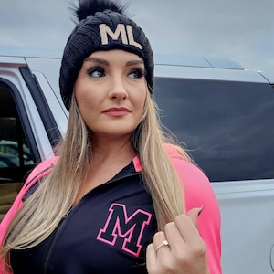 Puede incluir: Una chaqueta negra y rosa con cremallera que tiene una gran "M" blanca en la parte delantera. La chaqueta tiene rayas blancas y negras en las mangas y una boina negra con pompón con las letras "ML" en dorado en la parte delantera.