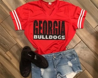 May include: Red and white jersey with the words "Georgia Bulldogs" in black and silver glitter. The jersey is paired with light blue denim shorts.