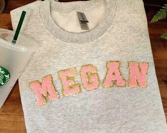 May include: A gray sweatshirt with the name "Megan" spelled out in pink and gold glitter letters.