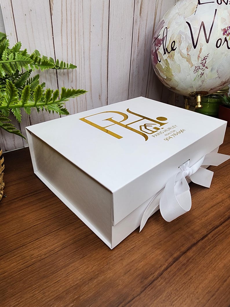 May include: A white gift box with a gold logo and text, tied with a white ribbon. The box is on a wooden surface. The logo reads "Paris Hurley Realtor" with a phone number.