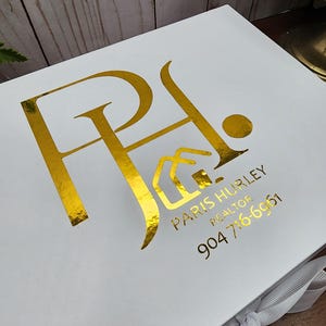 May include: White gift box with gold foil lettering and logo. The box features the initials "PA" with a house icon, the name "PARIS HURLEY REALTOR", and a phone number. A white ribbon is tied around the box.