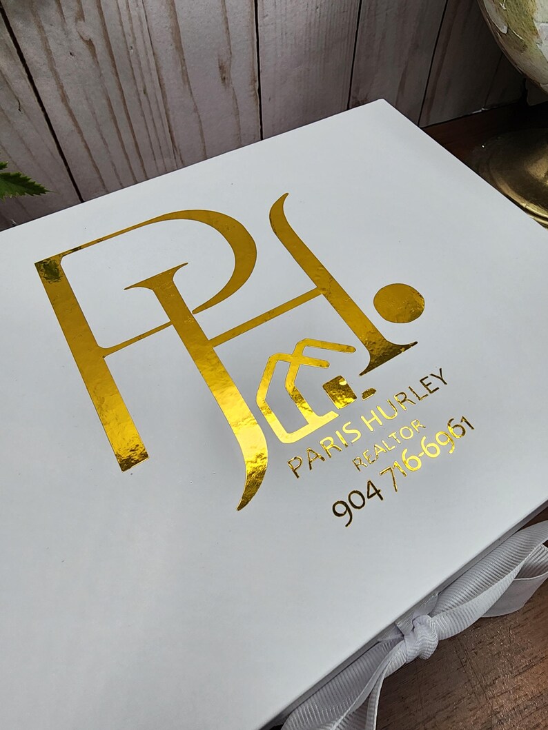 May include: White gift box with gold foil lettering and logo. The box features the initials "PA" with a house icon, the name "PARIS HURLEY REALTOR", and a phone number. A white ribbon is tied around the box.
