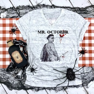 May include: White V-neck t-shirt with a distressed look. The shirt features a graphic of a man with the text "MR. OCTOBER" and "Michael Myers" printed on the front.