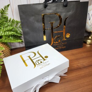 May include: A white gift box with a white ribbon and a black shopping bag. Both items have a gold logo that reads "Paris Hurley Realtor" and a phone number. The shopping bag has black handles.