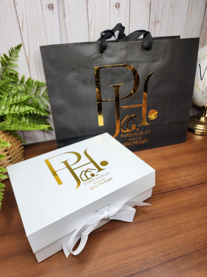 May include: A white gift box with a white ribbon and a black shopping bag. Both items have a gold logo that reads "Paris Hurley Realtor" and a phone number. The shopping bag has black handles.