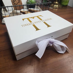 May include: A white gift box with gold lettering that reads "TT TINESHA TUTT REALTOR". The box is tied with a white ribbon. The box is on a wooden surface.